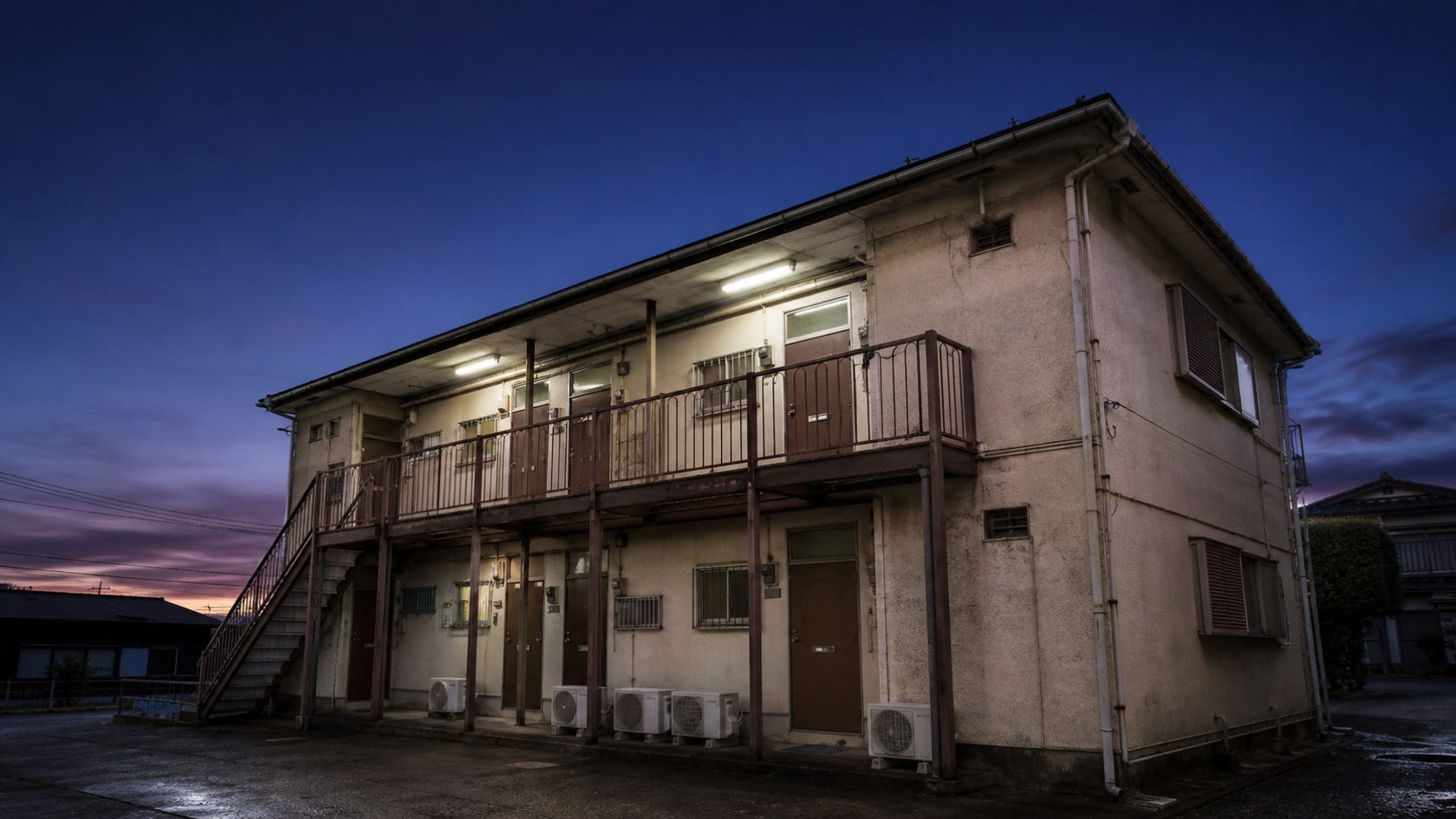夕暮れの古い木造アパート外観。外階段と並ぶ玄関ドア、共用廊下の蛍光灯が一部点灯し始めた佇まい。