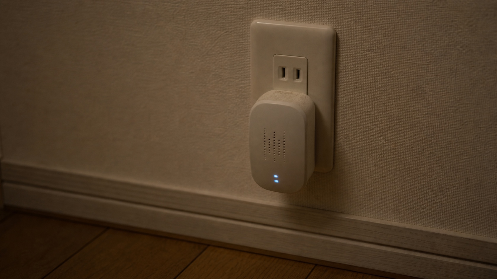 木造住宅の壁際コンセントに差し込まれた超音波害獣撃退器のクローズアップ。LEDインジケーターの微かな光が室内の薄暗がりに灯る情景。