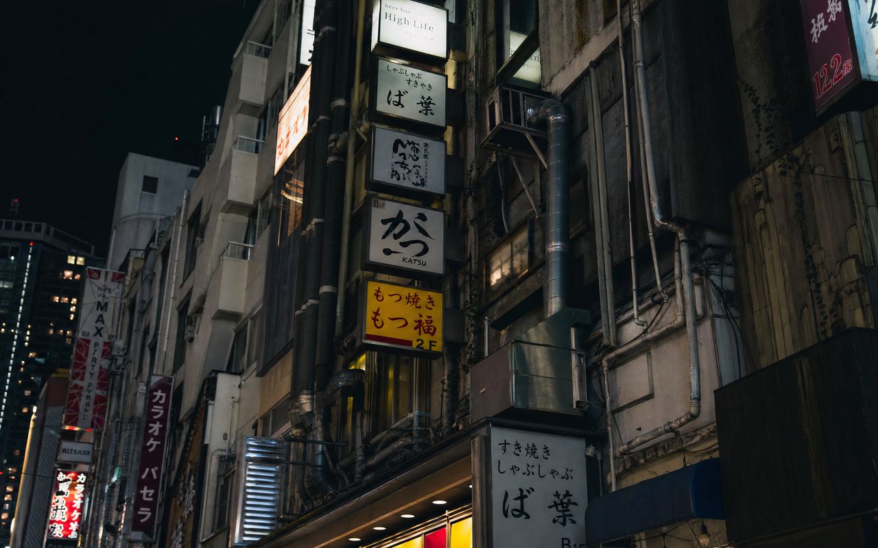 東京の繁華街にある築古飲食ビル。看板と外壁に這う配管が映る夜の街並みの写真。